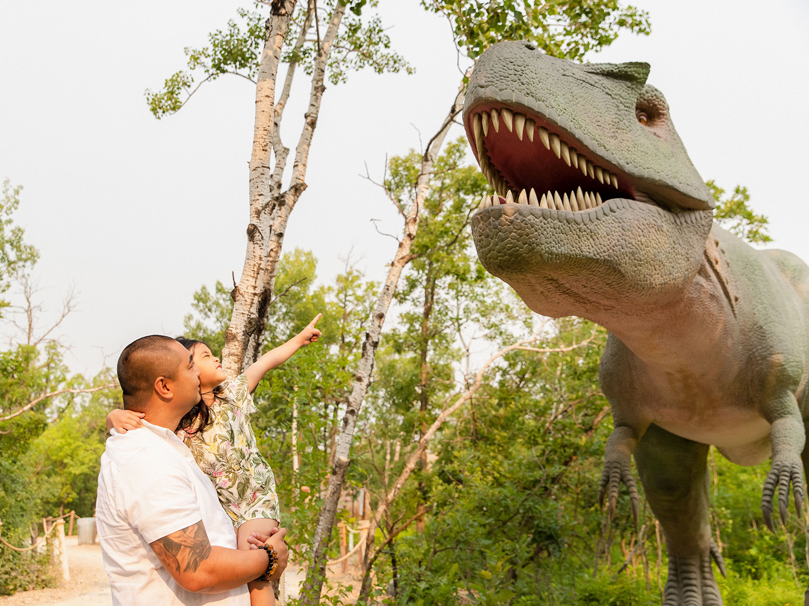 Father hold up young daughter who points at a large animatronic dinosaur
